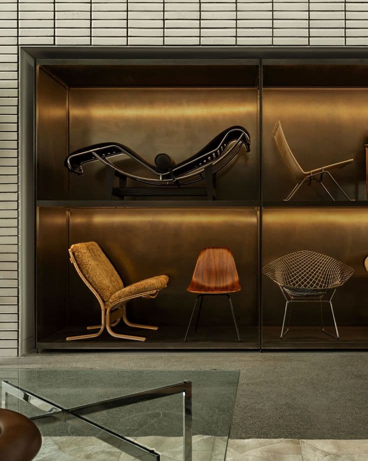 Iconic designer chairs displayed in a backlit bronze shelving unit against white brick.