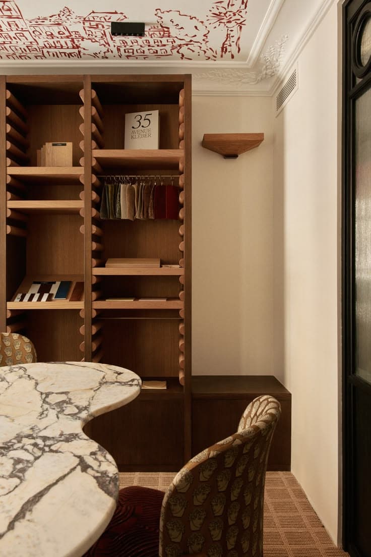 Interior design studio with marble table, patterned chairs, and wooden shelves displaying fabric samples.